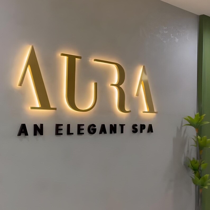 May include: The word "AURA" in gold, illuminated letters is mounted on a light gray wall. Below, the text "AN ELEGANT SPA" is in black letters. A green accent wall and small plant are visible.