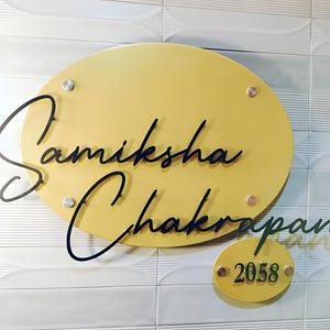 May include: A mustard yellow oval-shaped nameplate with the name "Samiksha Chakrapani" in black cursive script. A smaller, matching oval below displays the number "2058". The nameplate is mounted on a white tiled wall with silver and gold colored hardware.