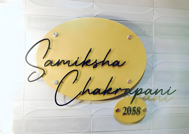 May include: A mustard yellow oval-shaped nameplate with the name "Samiksha Chakrapani" in black cursive script. A smaller, matching oval below displays the number "2058". The nameplate is mounted on a white tiled wall with silver and gold colored hardware.
