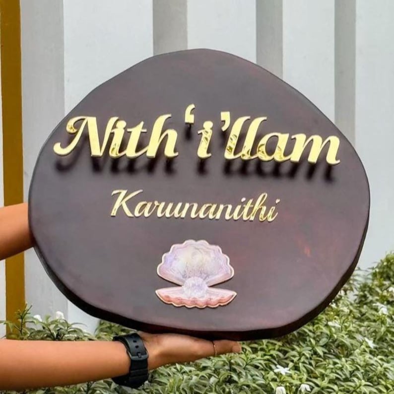 May include: A dark brown, oval-shaped wooden sign with gold lettering that reads "Nith'i'Ilam" and "Karunanithi". A seashell design is at the bottom. The sign is held in front of greenery.