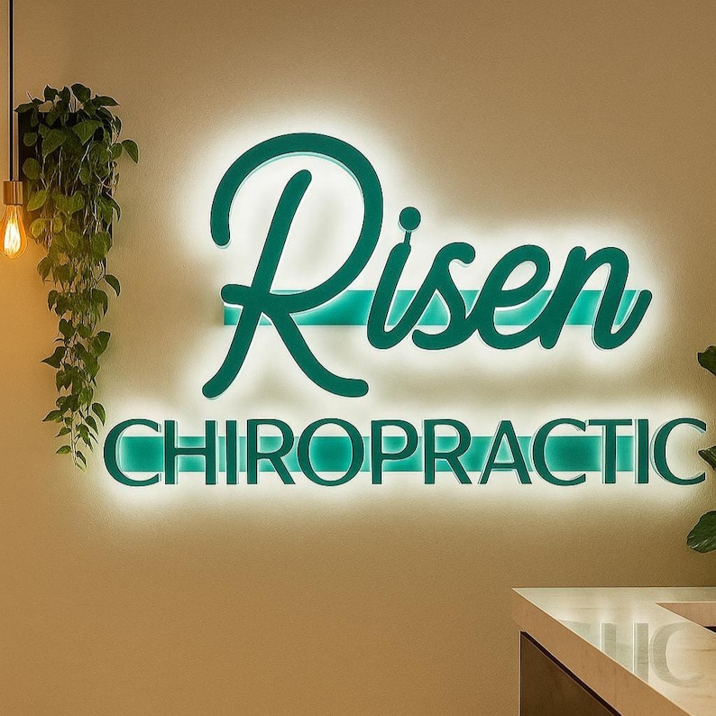 May include: A teal illuminated sign that reads "Risen Chiropractic" is mounted on a beige wall. The word "Risen" is in a cursive font, and "Chiropractic" is in a sans-serif font. A hanging plant and light fixture are visible.