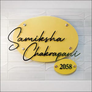 May include: A yellow oval-shaped nameplate with the name "Samiksha Chakrapani" in black cursive script. A smaller yellow oval below displays the number "2058". The nameplate is mounted on a white tiled wall with silver-colored screws.