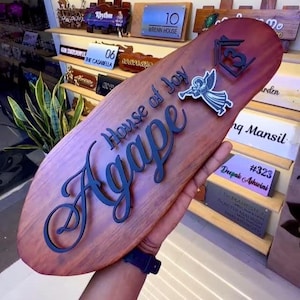 May include: A wooden sign with the words "House of Joy Agape" in a dark blue script. The sign has an oval shape and features an angel graphic and a house symbol. Other signs are visible in the background.