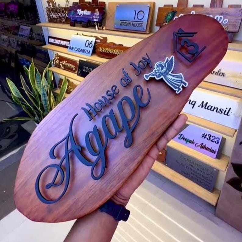May include: A wooden sign with the words "House of Joy Agape" in a dark blue script. The sign has an oval shape and features an angel graphic and a house symbol. Other signs are visible in the background.