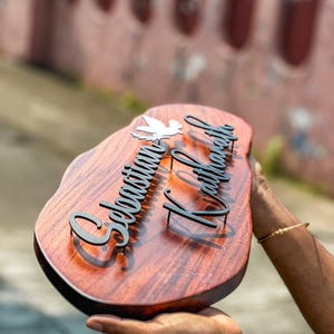 May include: A wooden nameplate with a dark brown finish, featuring the names "Sebastian Kaithara" in black, stylized lettering. A white dove is placed above the first name. The nameplate is held in a person's hands.