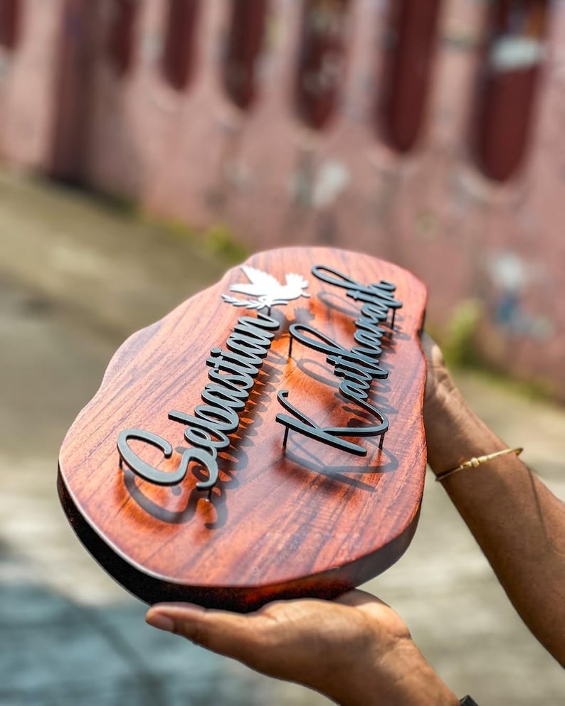 May include: A wooden nameplate with a dark brown finish, featuring the names "Sebastian Kaithara" in black, stylized lettering. A white dove is placed above the first name. The nameplate is held in a person's hands.