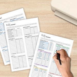 May include: Three sheets of white paper with a "Monthly Budget" template are displayed on a light wood surface. The template includes sections for income, expenses, and savings. A hand is writing on one sheet with a black pen. A printer is in the background.