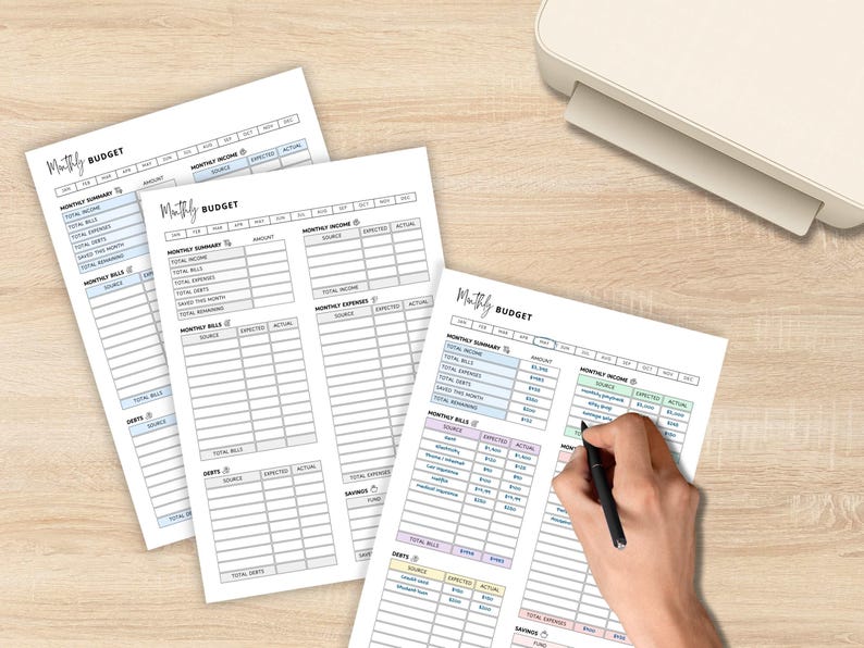 May include: Three sheets of white paper with a "Monthly Budget" template are displayed on a light wood surface. The template includes sections for income, expenses, and savings. A hand is writing on one sheet with a black pen. A printer is in the background.