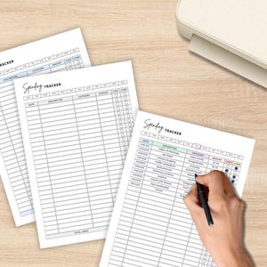 May include: Three white spending tracker sheets with a handwritten "Spending Tracker" title. One sheet has a hand filling it out with a black pen. A white printer sits in the background on a light wood surface.