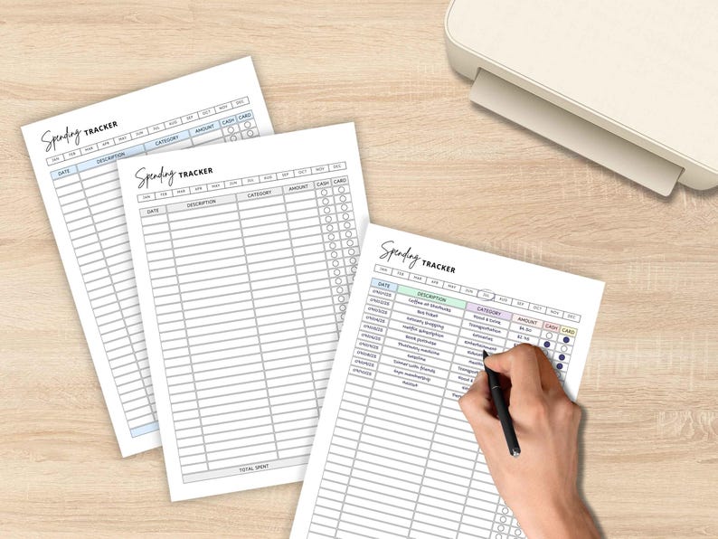 May include: Three white spending tracker sheets with a handwritten "Spending Tracker" title. One sheet has a hand filling it out with a black pen. A white printer sits in the background on a light wood surface.