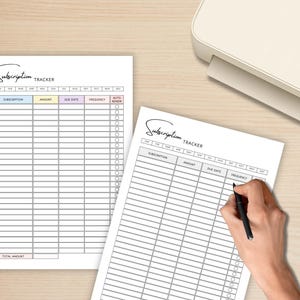 May include: Two white subscription tracker sheets with a light wood background. The sheets have a header that says "Subscription Tracker" and include columns for subscription, amount, due date, frequency, and auto-renew. A hand is holding a pen, writing on one of the sheets.