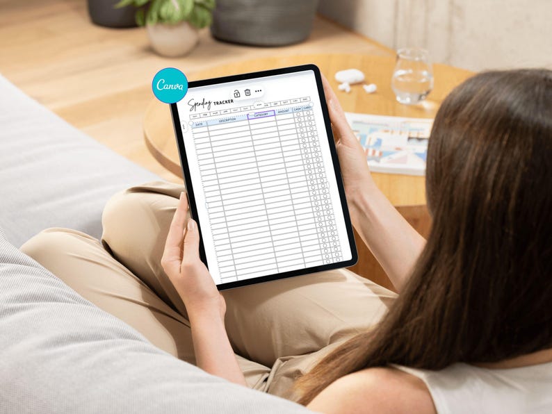 May include: A person holding a black tablet displaying a "Spending Tracker" template. The template has rows and columns for tracking expenses. The tablet is held in front of the person, who is sitting on a gray surface.