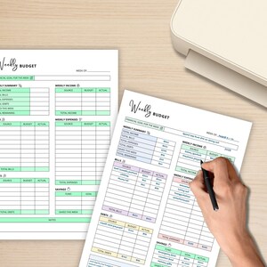 May include: Two white Weekly Budget sheets with pastel color-coded sections for income, expenses, bills, and savings. One sheet is being filled in with a black pen. The sheets are on a light wood-grain surface.