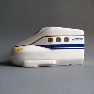 Puede incluir: Un tarro de galletas de cerámica blanca con forma de tren bala japonés con detalles azules y amarillos. La tapa está abierta y el tarro dice "N700" en el lateral.