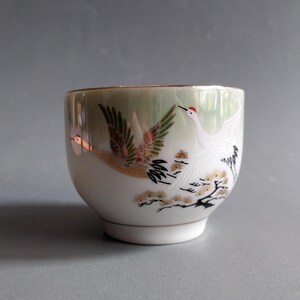 May include: A white ceramic teacup with a green glaze and gold accents. The cup features a painted design of two white cranes flying over a branch with white flowers.