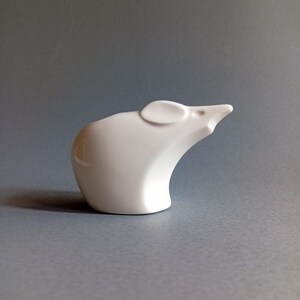 May include: A white ceramic mouse figurine with a smooth, glossy finish. The mouse has a curved body, a small ear, and a pointed snout. The figurine is set against a gray background, highlighting its minimalist design.