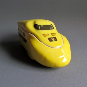 Puede incluir: Una caja de cerámica amarilla en forma de tren bala japonés. La caja tiene una franja negra y detalles negros en la parte delantera, incluido el texto "T5".