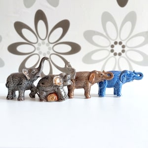 May include: Five ceramic elephant figurines in various colours, including grey, brown, and blue. The elephants are arranged in a row, facing the viewer.