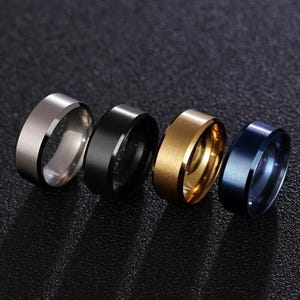 May include: Four metal rings in a row, each in a different colour. The rings are silver, black, gold, and blue. The rings have a smooth, polished finish and are all the same width. The rings are displayed on a dark surface.