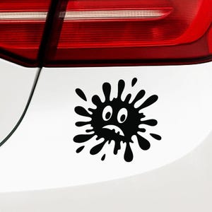 May include: Black vinyl car sticker featuring a cartoon splatter design with a sad face. The sticker is applied to a white car surface near the rear light. The splatter has a unique, irregular shape.