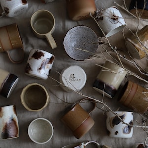 May include: A collection of handcrafted ceramic mugs, bowls, and a small container with the word "POT" visible. The mugs display diverse designs, including speckled, striped, and solid colours in shades of brown, beige, and white. The items are arranged on a neutral-toned surface.