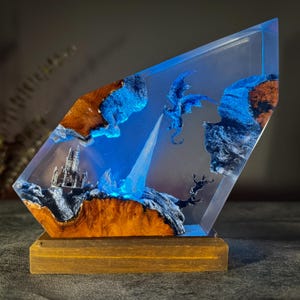 May include: A fantasy-themed resin sculpture featuring a blue dragon, a castle, and a waterfall. The sculpture is encased in clear resin with blue and brown accents, mounted on a wooden base. The dragon is mid-flight, with a waterfall flowing from the top.