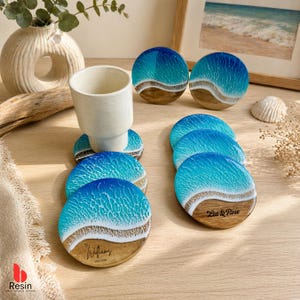 Puede incluir: Un juego de posavasos redondos de resina con diseño de playa. Los posavasos presentan un patrón de olas azules y blancas sobre una base color arena. Algunos posavasos tienen texto personalizado. Una taza de cerámica blanca descansa sobre un posavasos.