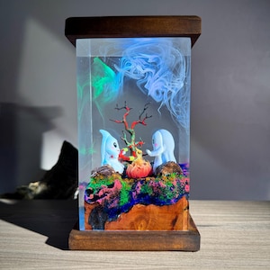 May include: A decorative resin lamp with a fantasy underwater scene. Two blue cartoon figures stand near a red and orange tree, set against a backdrop of swirling blue and green colours. The lamp has a wooden base and top.