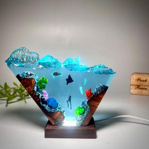 May include: A unique resin lamp featuring an underwater scene with a diver, manta ray, and coral. The lamp has a wooden base and a clear, diamond-shaped resin body with blue and turquoise accents. The lamp is lit from the base.
