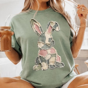 Peut inclure: T-shirt vert sauge avec un motif de lapin en patchwork rose, bleu et crème. Le lapin a des bords effilochés. Le t-shirt est de coupe décontractée. La personne porte un short blanc et des chaussettes à rayures noires. On voit aussi un verre de café glacé.
