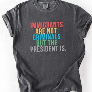 May include: Dark gray t-shirt with the text "IMMIGRANTS ARE NOT CRIMINALS BUT THE PRESIDENT IS." in red, blue, green, and gray. The shirt has a crew neck and short sleeves. The fabric appears to be pre-washed.
