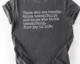 May include: Dark gray t-shirt with rolled sleeves. The shirt features white text that reads: "Those who see beauty, think beautifully; and those who think beautifully, find joy in life."