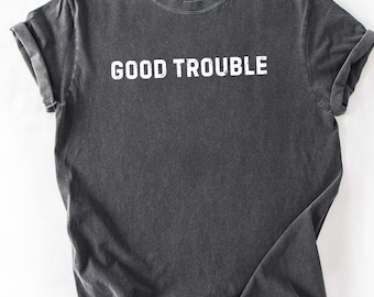 May include: A dark gray t-shirt with the words "GOOD TROUBLE" printed in white block letters. The shirt has a crew neck and short sleeves, with the sleeves rolled up. The fabric appears soft and slightly worn.