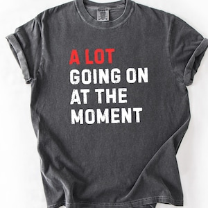 May include: Dark gray t-shirt with the phrase "A LOT GOING ON AT THE MOMENT" printed in white and red. The shirt has a crew neck and short sleeves, with a slightly faded, vintage look.