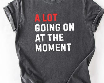 May include: Dark gray t-shirt with the phrase "A LOT GOING ON AT THE MOMENT" printed in white and red. The shirt has a crew neck and short sleeves, with a slightly faded, vintage look.