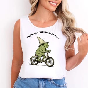 May include: White vest top with the text "Off to commit mom foolery." The design features a cartoon frog wearing a hat and sunglasses, riding a bicycle. The frog is green, and the bicycle is white.