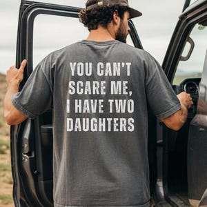 May include: Gray t-shirt with the text "YOU CAN'T SCARE ME, I HAVE TWO DAUGHTERS" in white. The person is wearing a brown cap and dark jeans, standing next to a vehicle with the door open.