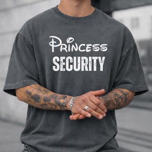 May include: Dark gray t-shirt with the text "Princess Security" in white. The word "Princess" is written in a stylized font. The shirt has short sleeves and a crew neck.
