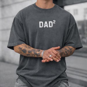 May include: Dark gray t-shirt with the text "DAD²" in white, featuring a crew neck and short sleeves. The shirt has a slightly faded appearance. The image shows the shirt on a person.
