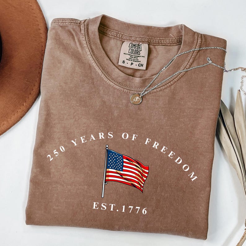 Pu&ograve; includere: Una maglietta Comfort Colors marrone con la scritta "250 YEARS OF FREEDOM" e una grafica della bandiera americana. La maglietta ha anche la scritta "EST. 1776". Una collana d'argento con un ciondolo &egrave; sulla maglietta.