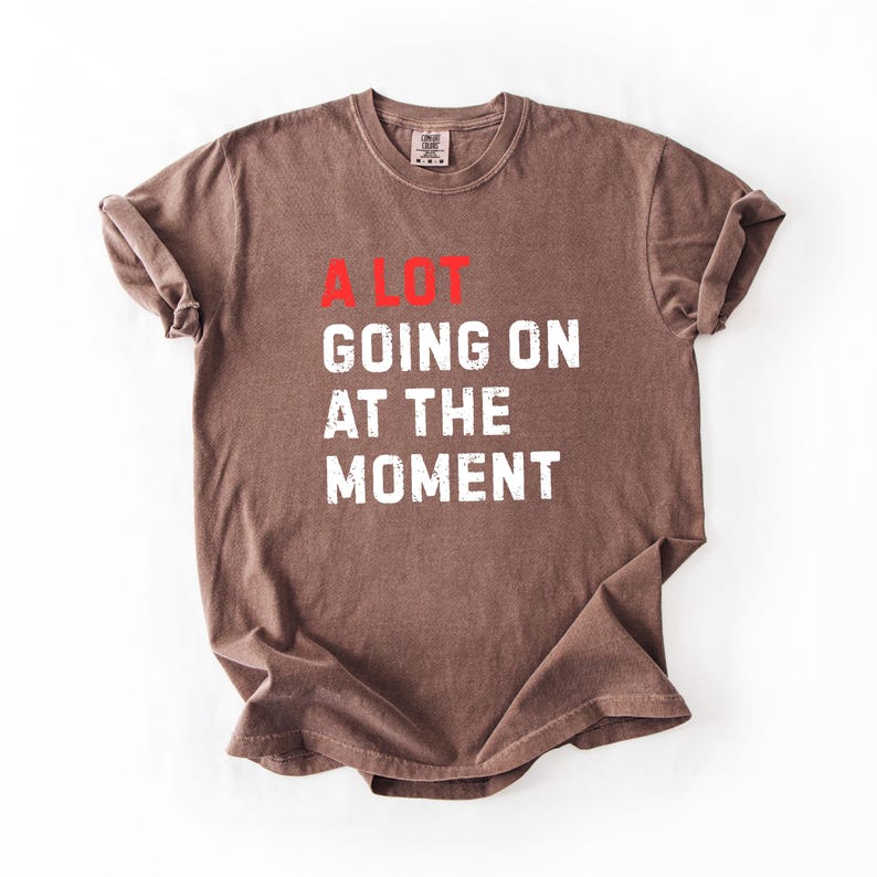 May include: Brown t-shirt with the text "A LOT GOING ON AT THE MOMENT" in white and red. The shirt is folded and laid flat on a white surface. The shirt has a crew neck and short sleeves.