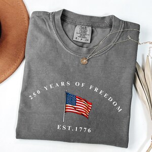 Pu&ograve; includere: Una maglietta grigia con la scritta "250 YEARS OF FREEDOM" e un'illustrazione della bandiera americana. La maglietta ha anche la scritta "EST. 1776". Una collana d'argento con un ciondolo &egrave; sulla maglietta.