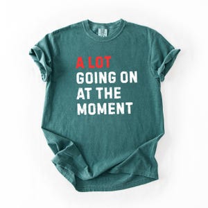 May include: A teal-colored t-shirt with the phrase "A LOT GOING ON AT THE MOMENT" printed in white block letters. The word "LOT" is in red. The shirt has a crew neck and short sleeves.