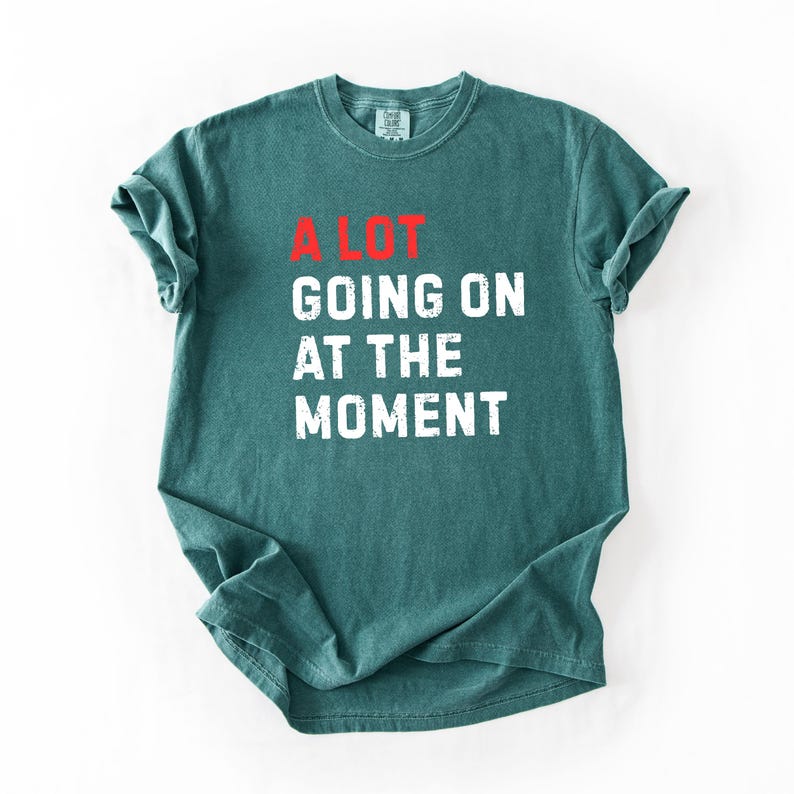 May include: A teal-colored t-shirt with the phrase "A LOT GOING ON AT THE MOMENT" printed in white block letters. The word "LOT" is in red. The shirt has a crew neck and short sleeves.