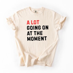 May include: Cream-colored t-shirt with the phrase "A LOT GOING ON AT THE MOMENT" printed in red and black. The shirt has a classic crew neck and short sleeves.