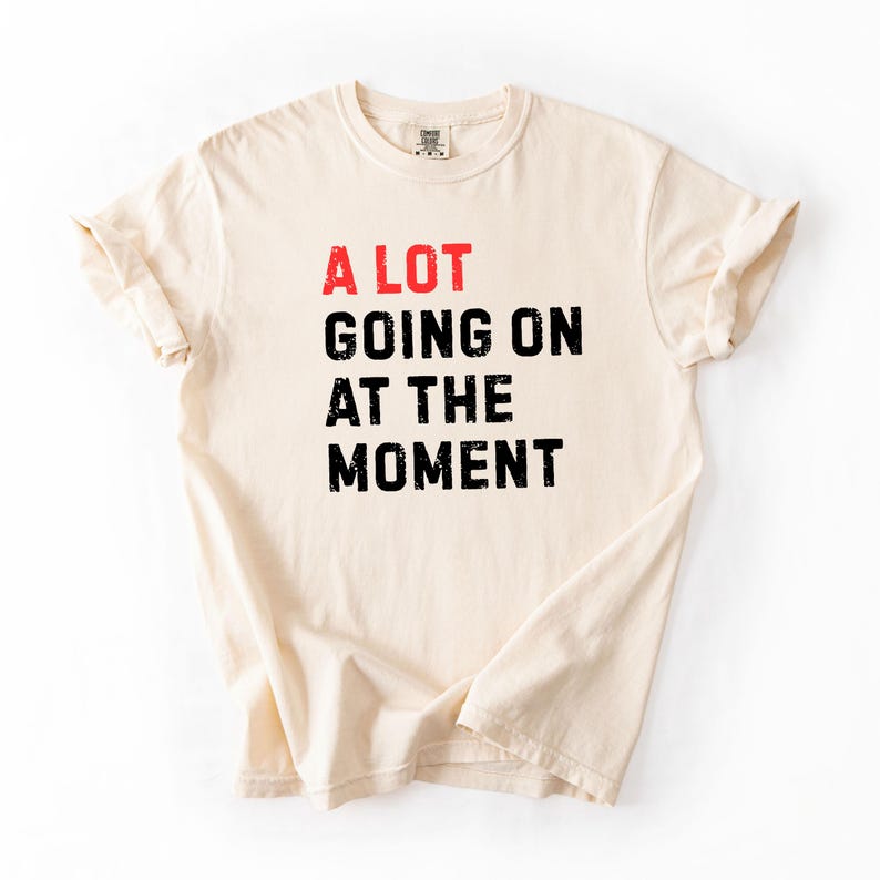May include: Cream-colored t-shirt with the phrase "A LOT GOING ON AT THE MOMENT" printed in red and black. The shirt has a classic crew neck and short sleeves.