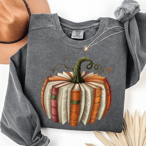 May include: Dark gray sweatshirt featuring a pumpkin design made of open books. The pumpkin has an orange and white color scheme with a green stem and vines. A brown hat and necklace are visible in the background.