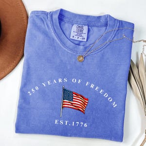 Pu&ograve; includere: Una maglietta blu Comfort Colors con la scritta "250 YEARS OF FREEDOM" e "EST. 1776" che circondano una bandiera americana. Una collana d'oro con un ciondolo &egrave; drappeggiata sulla maglietta.