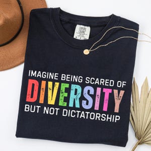 May include: A navy blue t-shirt with the text "IMAGINE BEING SCARED OF DIVERSITY BUT NOT DICTATORSHIP" in rainbow colors. The shirt has a Comfort Colors tag and is paired with a gold necklace and a brown hat.