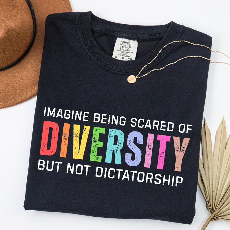 May include: A navy blue t-shirt with the text "IMAGINE BEING SCARED OF DIVERSITY BUT NOT DICTATORSHIP" in rainbow colors. The shirt has a Comfort Colors tag and is paired with a gold necklace and a brown hat.